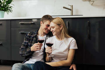 Happy couple drinking wine while sitting on a floor in the kitchen. They enjoy romantic time on family weekend together.