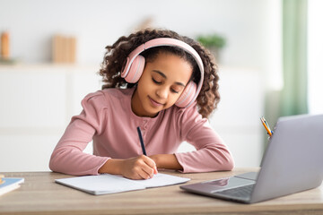 African american schoolgirl having online lesson, using laptop and headphones at home, taking notes