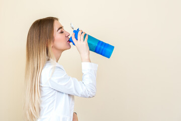 Side view of beautiful young caucasian woman is drinking water from plastic bottle against a beige background
