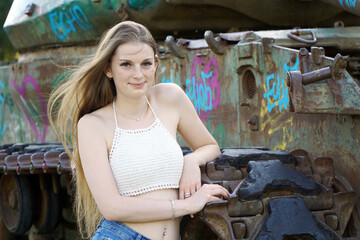 Pretty young woman wears hot pants and top bellyless, leaning against old tank and smiling friendly...