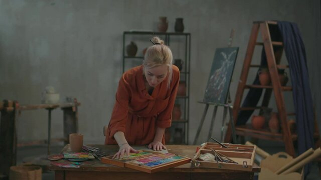 Professional charming female artist choosing colors from set of multicolored pastel crayons in open wooden artist box on rustic table, drawing sketch on paper while creating in art workshop.