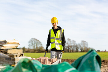 A young adult male builder wearing a high visibility vest and hard hat pushing a wheelbarrow full of bricks while on a building site