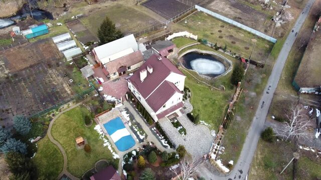 Aerial view of a country house. The plot has a garden with trees and conifers, a swimming pool, a stable and outbuildings surrounded by winter nature. Ukraine, Poltava. February 2021