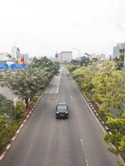 A beautiful black car crosses the middle of Jakarta Indonesia's road, on the left and right of the road, there are shady trees, making the road more comfortable for vehicles to pass.