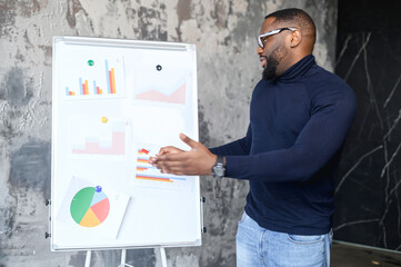 Confident African-American male employee points on whiteboard with graphs, planning strategy, smart and intelligent freelancer guy presenting new project, sturtup idea, office employee reports