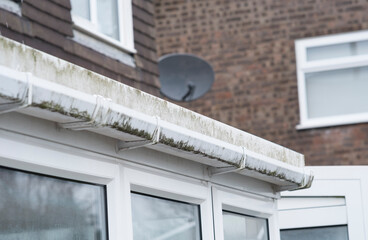 Dirty clogged white plastic pvc gutters and drain pipes with mossy green mould on plastic fascias. ...
