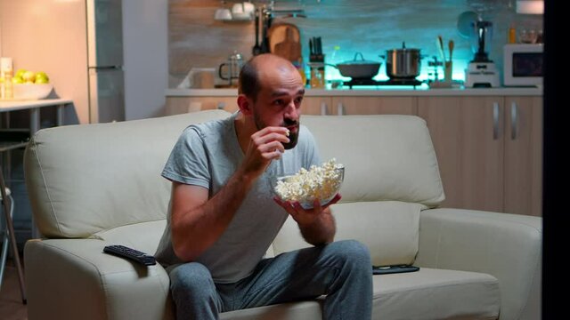 Upset caucasian man watching sport match alone in the living room while hus favourite team looses the game, eating popcorn late at night