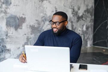 Successful African-American male employee using the laptop for work at modern loft office, watching webinars and taking notes with a pen, working on a new project, businessman plans tasks for the week