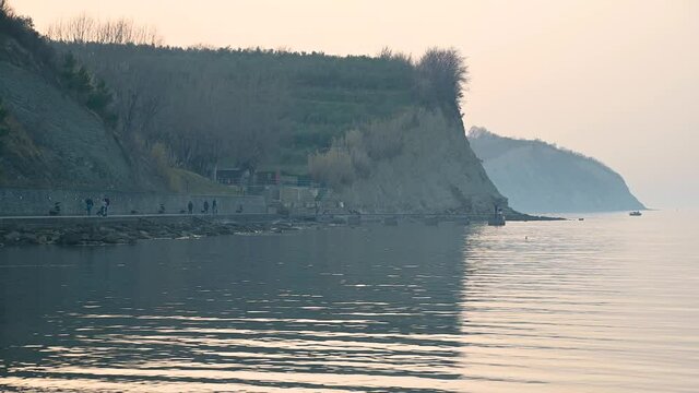 San Simon bay, Slovenia: people walking on walkway shore in winter evening. Calm waves. Beautiful Adriatic Sea. Sunset behind famous cliffs. Static shot, real time