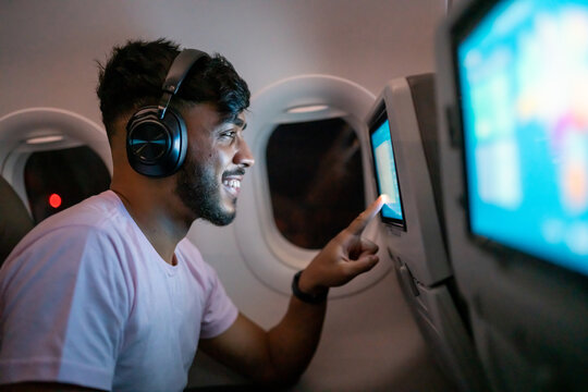 Passenger In Airplane Touching LCD Entertainment Screen. Latin American Man In Plane Cabin Using Smart Device Listening To Music On Headphones.