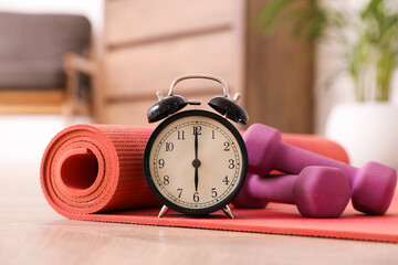 Alarm clock, yoga mat and dumbbells on wooden floor indoors. Morning exercise