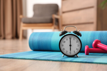 Alarm clock, yoga mat and dumbbells on wooden floor indoors. Morning exercise