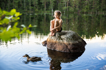 Cute little girl sitting on a rock in lake. Enjoying summer vacation. Child and Nature. Happy isolation concept. Exploring Finland. Scandinavian landscape. 