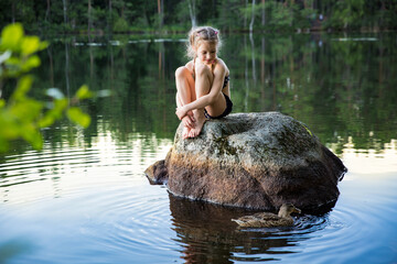 Cute little girl sitting on a rock in lake. Enjoying summer vacation. Child and Nature. Happy isolation concept. Exploring Finland. Scandinavian landscape. 