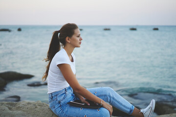 Fototapeta na wymiar Pondering hipster girl dressed in casual jeans holding sketchbook for training draw skills and thoughtful looking at seascape, pensive female student with education textbook thinking about knowledge