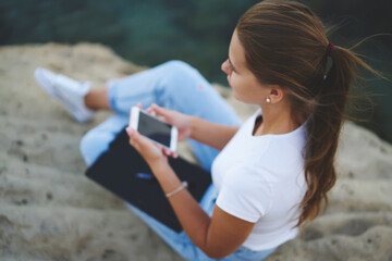 Top view of millennial hipster girl with modern smartphone technology pondering on text publication, teenage female blogger with mobile technology and sketchbook for learning resting outdoors