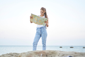 Caucasian female tourist reading location paper map search orientation outdoors, attractive hipster girl in denim jeans 20 years old exploring seashore area during travel weekend or vacations
