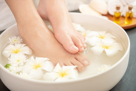 Closeup View Of Woman Soaking Her Feet In Dish With Water And Flowers On Wooden Floor. Spa Treatment And Product For Female Feet And Hand Spa. White Flowers In Ceramic Bowl.