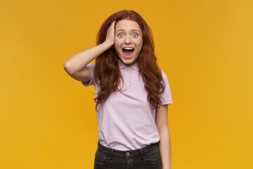 Fototapeta premium Shocked, positive woman with long ginger hair. Wearing pink t-shirt. People and emotion concept. Touching her head, surprised. Watching at the camera, isolated over orange background