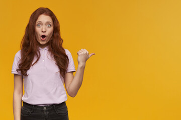 Shocked girl, surprised redhead woman with long hair. Wearing pink t-shirt. Emotion concept. Pointing thumb to the right at copy space and watching at the camera, isolated over orange background