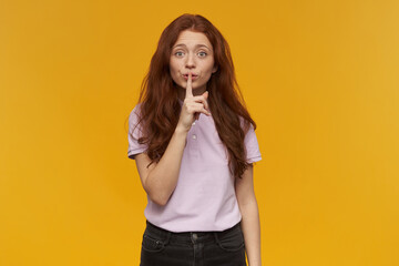 Cute, attractive woman with long ginger hair. Wearing pink t-shirt. People and emotion concept. Showing silence sign, asks to be quiet. Watching at the camera, isolated over orange background