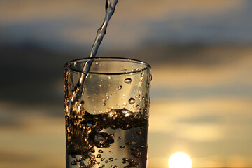 Pouring clean water into drinking glass on evening sun background. Concept of health and freshness,...
