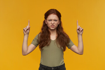 Negative, frowning woman with long ginger hair. Wearing green t-shirt. People and emotion concept. Watching at the camera and pointing up at copy space, isolated over orange background