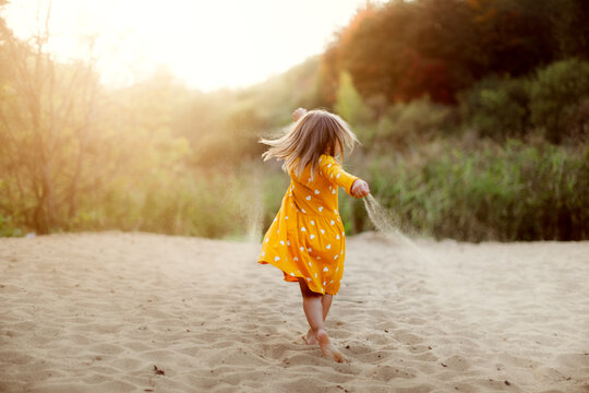 Caucasian cute little girl in yellow dress dancing on the sand in summer at sunset, happy childhood and freedom