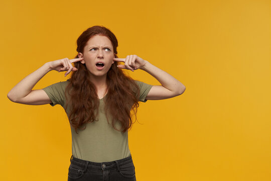 Irritated looking girl, frowning redhead woman with long hair. Wearing green t-shirt. Emotion concept. Close her ears with fingers. Watching to the right at copy space, isolated over orange background
