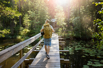 Mature man exploring Finnish nature in summer, walking across the bridge. Hiker with big backpack...