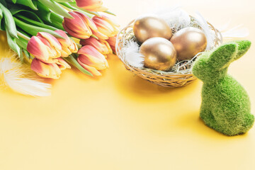 Easter eggs basket. Golden egg in basket with spring tulips, white feathers on pastel yellow background in Happy Easter decoration. Traditional decoration in sun light.