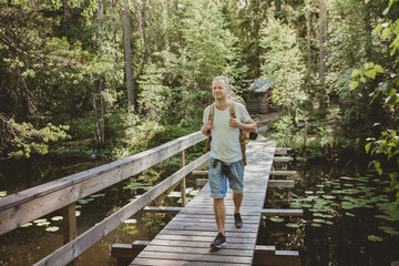 Fototapeta na wymiar Mature man exploring Finnish nature in summer, walking across the bridge. Hiker with big backpack traveling in forests. Summer Scandinavian landscape of lakes and woods. 