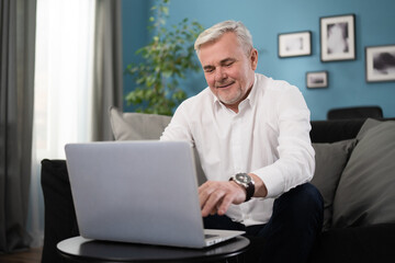 Fototapeta premium Smiling happy middle aged 50s old senior man using laptop computer technology working online, surfing internet, typing email, shopping in ecommerce store relaxing with device sitting on couch at home.