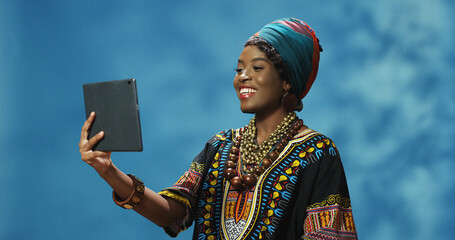 African American woman in traditional outfit holding tablet device and talking via webcam....