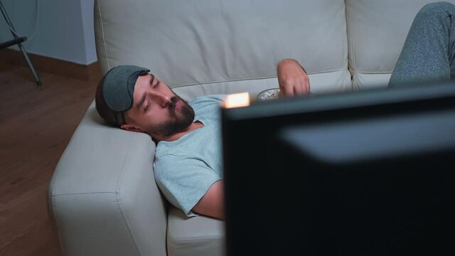 Caucasian male with eye sleep mask eating popcorn while watching entertainment tv show. Tired man sitting in front of television during movie film late at night in kitchen