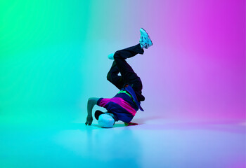 Beautiful sportive boy dancing hip-hop in stylish clothes on colorful gradient background at dance hall in neon light.
