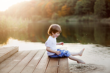 Fototapeta premium Caucasian cute boy on the footbridge by the lake, happy childhood in the village, child play in the park near the water, swing their feet in the water of the lake