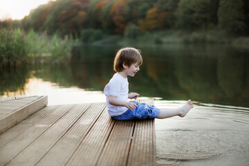 Caucasian cute boy on the footbridge by the lake, happy childhood in the village, child play in the park near the water, swing their feet in the water of the lake