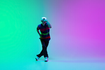 Beautiful sportive boy dancing hip-hop in stylish clothes on colorful gradient background at dance hall in neon light.