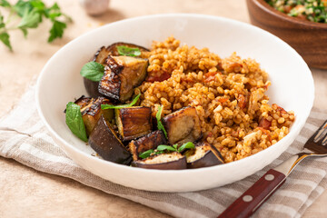 Bulgur pilaf (pilavi) with tomatoes and fried eggplants in a white plate on a light table close-up. Turkish cuisine, vegan food.