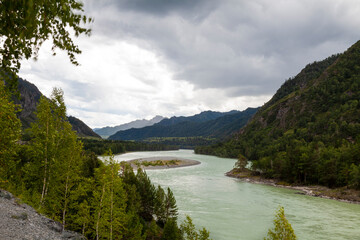 The bank of the Katun river with water containing turquoise clay and a sandy beach, rocky coast against the backdrop of mountains covered with green forest and blue sky with white clouds.