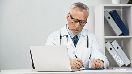 Fototapeta premium serious doctor writing diagnosis on clipboard near laptop at workplace