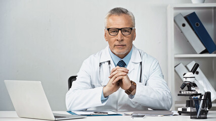 doctor in eyeglasses sitting near laptop and looking at camera