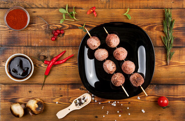 Meat balls on wooden skewers lie on a black plate. Sauce, spices, pepper pods, mushrooms, rosemary.