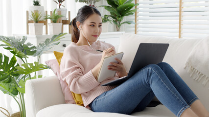 Cozy relax young attractive asian female employee wear earphone listen to digital online course development program in quarantine social distance at home reskill upskill school class for career path.