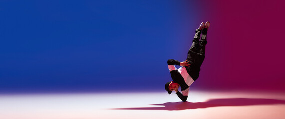 Beautiful sportive boy dancing hip-hop in stylish clothes on colorful gradient background at dance hall in neon light.