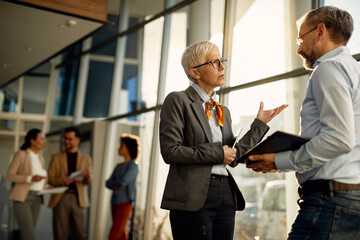 Mature businesswoman talking to male co-worker in hallway of office building.