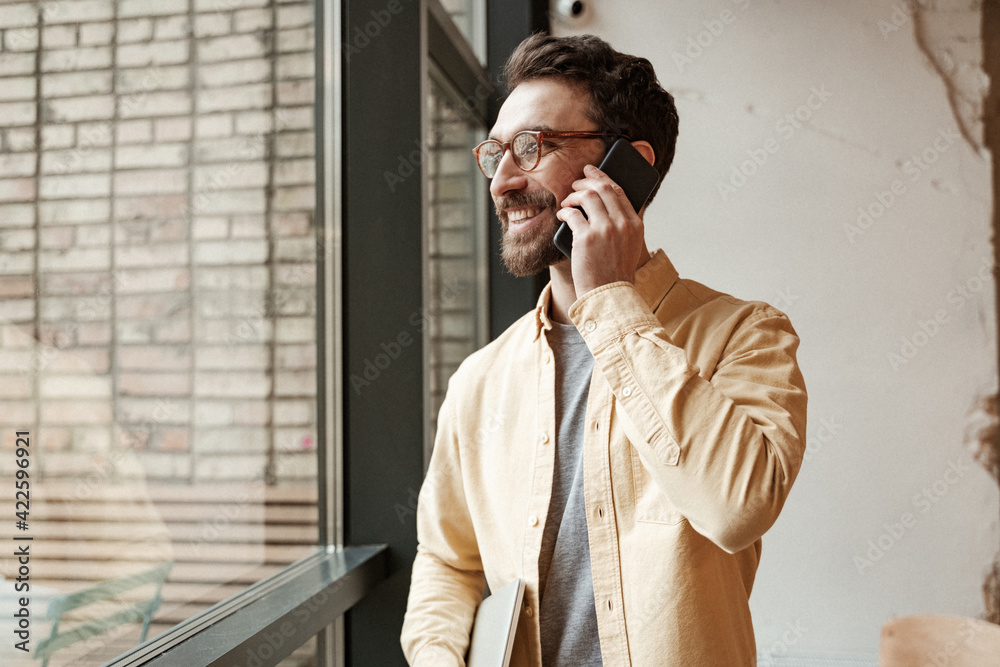 Wall mural happy and bearded freelancer holding laptop while standing near window and talking on smartphone