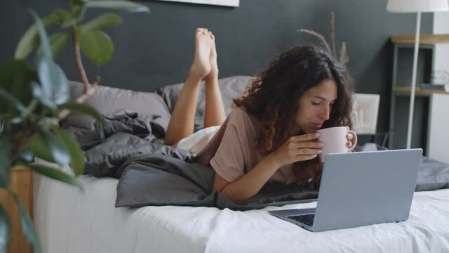 Young pretty woman lying on stomach on bed, drinking coffee, smiling and using laptop at home