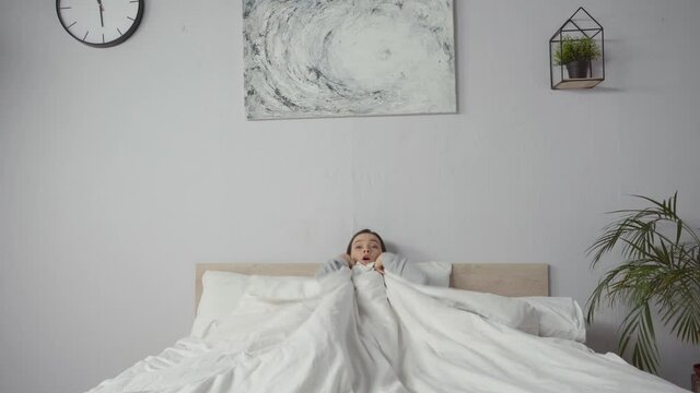 young woman jumping into bed and covering with blanket in bedroom
